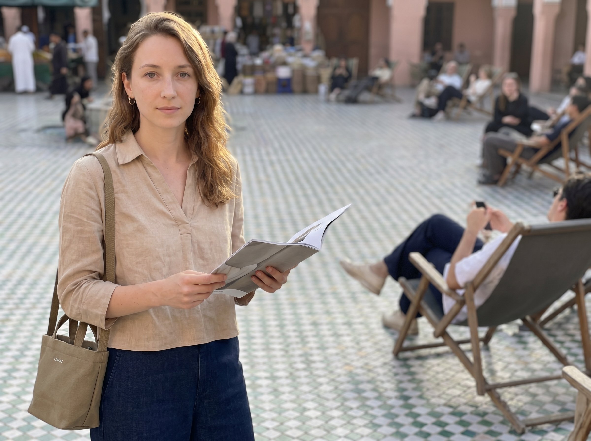 Three-quarter standing at the COMMUNE-Aoyama courtyard-corner-bank Sunday late-afternoon 16:45 JST holding a creased Lemaire SS27 lookbook open in the left hand plus a small khaki-canvas Lemaire-Tokyo-Showroom-tote slung over the right shoulder, 4:3
