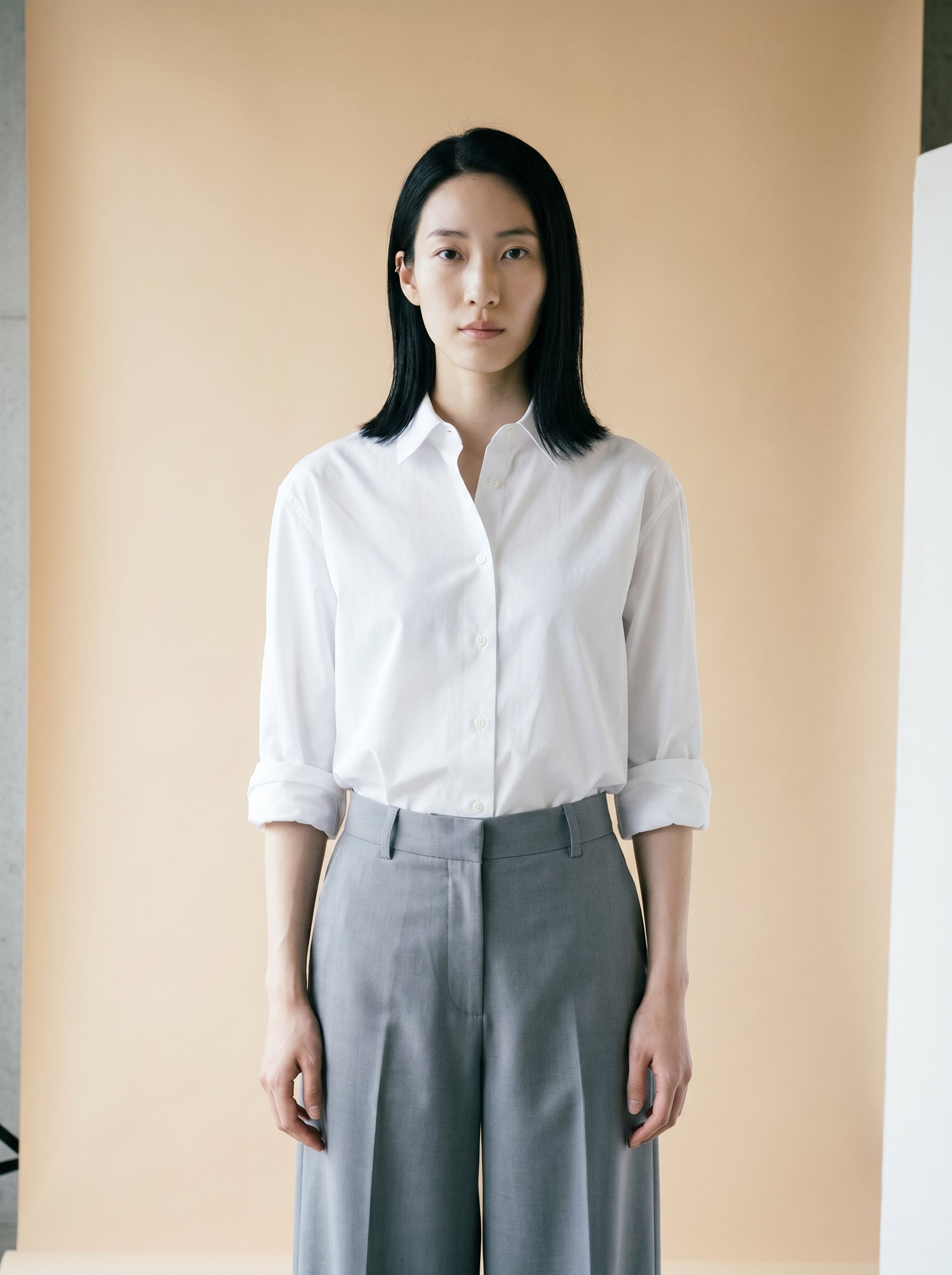 Final 1 — Hana on the soft-beige paper-roll backdrop, white poplin shirt and wide dove-grey tailored trouser, direct gaze