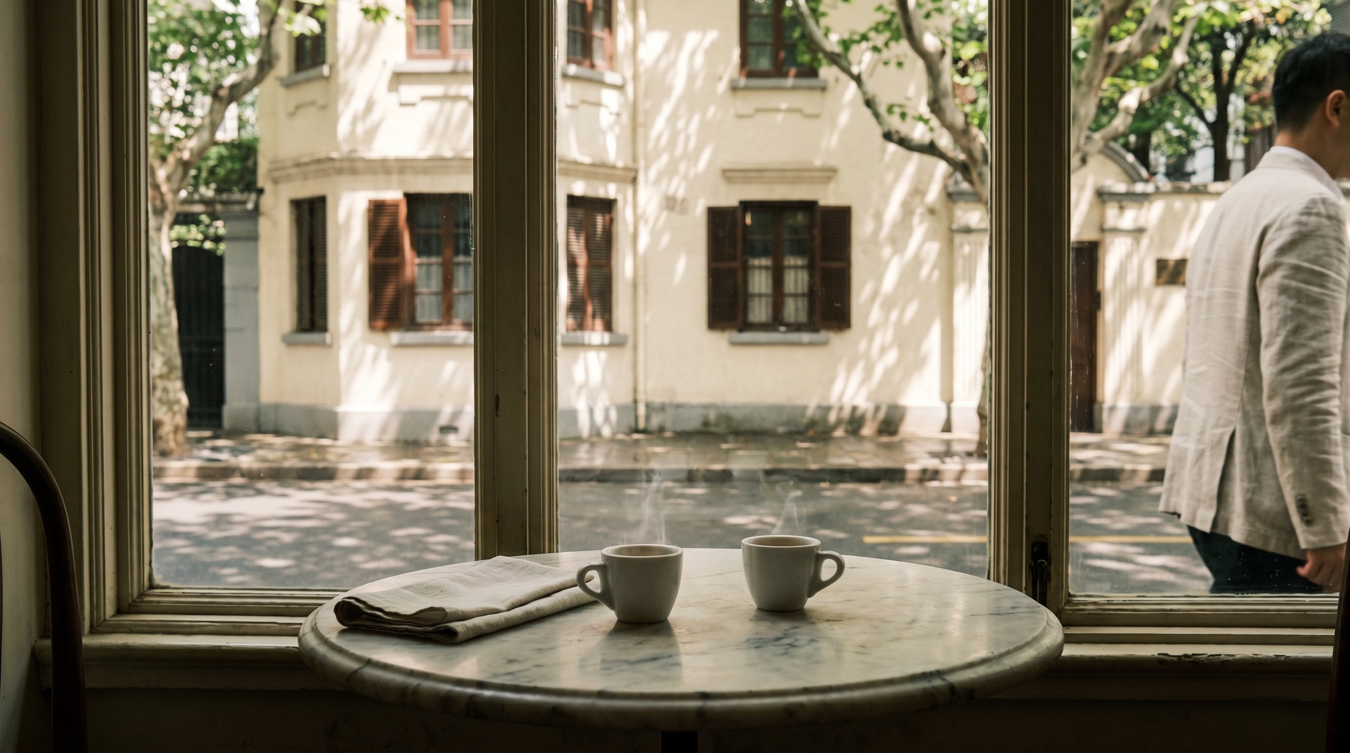 Anfu Lu Monday lunch hour, Egg cafe corner, two espresso cups on a small marble round-table, soft window-light on the wagner-villa facade behind, a single linen-shouldered figure half-out of frame